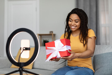 Happy black woman holding gift box and shooting video for blog, looking at cellphone set on ring lamp tripod