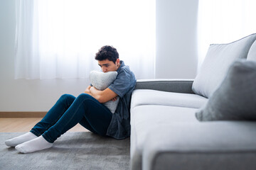 Pensive sad hispanic teenager boy sitting on floor and hugging pillow. Depression, anxiety and mental health concept - obrazy, fototapety, plakaty