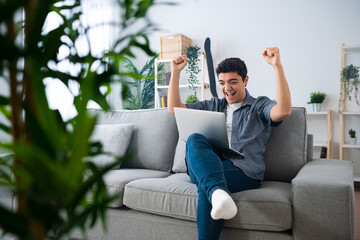 Teenager boy celebrating success in front of laptop while sitting on couch