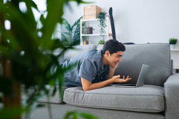 Hispanic teenager boy lying down on couch and talking on laptop. Live streaming and learning from home concept