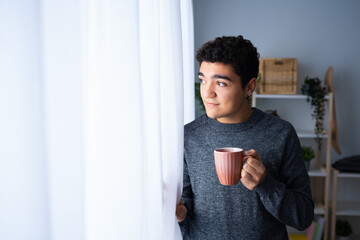 Portrait of confident hispanic teenager boy looking through the window and holding cup of coffee