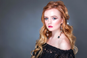 A beautiful red-haired girl with wavy hair, makeup, bare shoulders and long earrings posing on a gray background in the studio. woman looking at camera