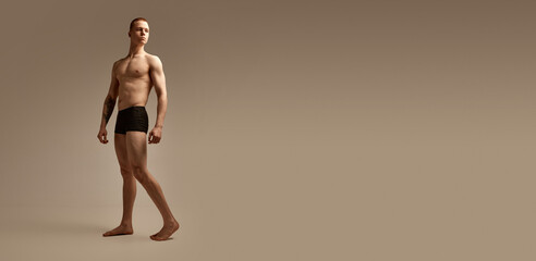 Full-length portrait of young man with fit, strong, muscular body posing in black boxers on studio...