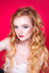 A beautiful red-haired girl with wavy hair, make-up, in a silk top and long earrings poses on a red background in the studio. woman is looking at camera