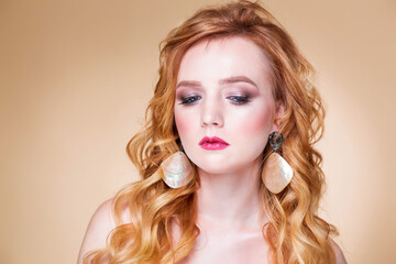Fototapeta premium Beautiful red-haired girl with wavy hair and makeup posing on a beige background in the studio in large gold earrings