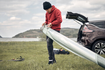 Windsurfer and camper unpacking equipment from a car in nature near the lake shore.