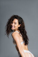 Fototapeta premium Cheerful curly woman in lingerie looking at camera isolated on grey.