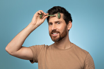 Portrait of handsome man using face roller for smooth and firm anti aging skin treatment, blue background