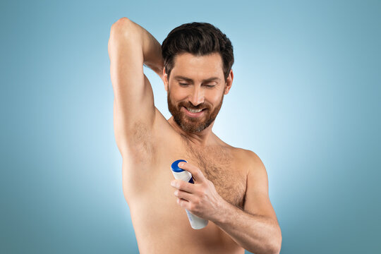 Shirtless caucasian man putting deodorant underarm after shower, standing over blue background, studio shot, free space