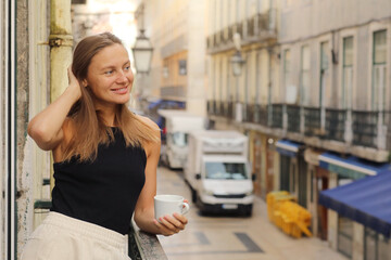 Happy young beautiful woman holds cup of coffee or tea, standing on the balcony with Europe city street in the morning. Mature female enjoying vacation in resort. Summer, weekend, tourism, travel