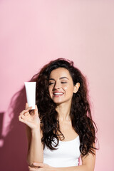 Naklejka premium Cheerful freckled woman looking at cosmetic cream on pink background.