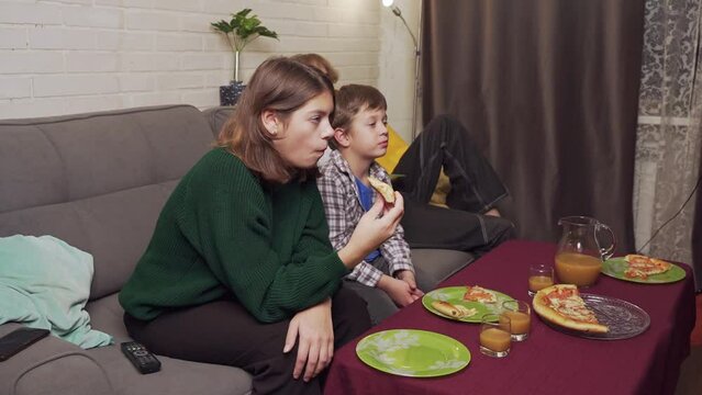 children are sitting at home on the couch, eating pizza, watching TV
