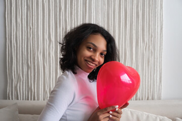 Closeup portrait of Young african american woman holding red heart shaped balloon sitting on sofa at home. Celebrating valentines,love day, women's day, birthday. Copy space banner
