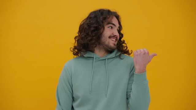 Smiling long-haired man in a blue hoodie talking at the camera and pointing to the copyspace in the yellow studio