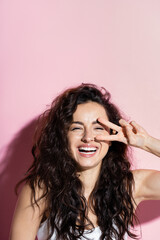 Obraz premium Cheerful curly woman showing victory sign on pink background.