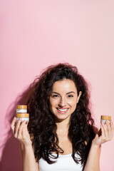 Curly young woman holding cosmetic creams on pink background.