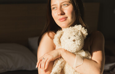 Portrait of girl is waking up in morning stretches in bed and sun shines from window. Close-up Happy young woman greets new sunny day and holds teddy bear toy