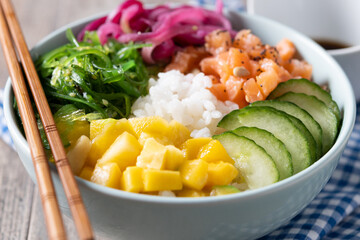 Poke bowl with rice, salmon,cucumber,mango,onion,wakame salad, poppy seeds ands sunflowers seeds on wooden background. Close up
