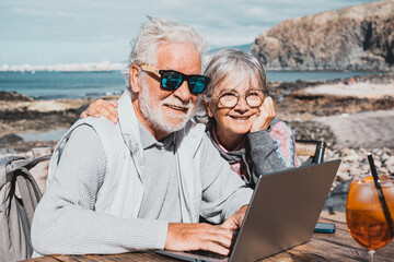 Cheerful attractive senior couple browsing together on laptop enjoying sunny day outdoor at the beach sitting at bar wooden table with orange cocktail. Elderly people in vacation or retirement at sea