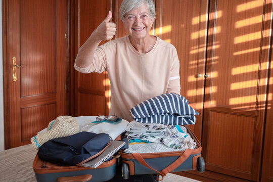 Happy senior woman thumb up looking into the camera while packing her clothes in a suitcase preparing for the summer vacation trip. Travel preparation concept