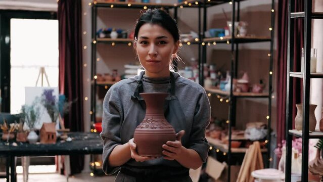A young woman potter holding a vase of clay. Brown vase from clay in the female potter's hands. The potter works in a pottery workshop with clay