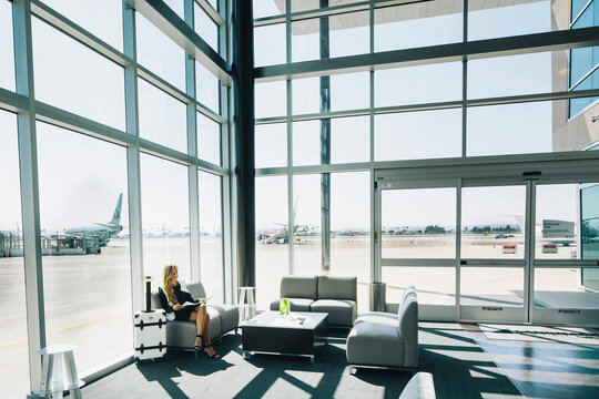 A beautiful lady sits on a sofa in a large glass fitted airport terminal.