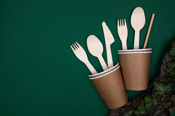 Disposable bamboo cutlery in a paper cup on a dark green background. Flat lay, top view, copy space.