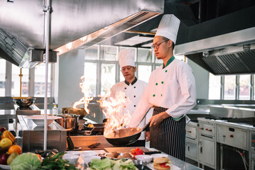 Professional mature chef looking to young chef cooking foods with high burning flames in restaurant.