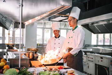 Professional mature chef looking to young chef cooking foods with high burning flames in restaurant.