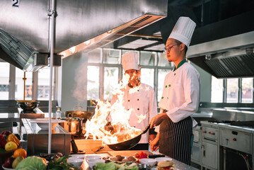 Professional mature chef looking to young chef cooking foods with high burning flames in restaurant.