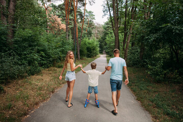 Fototapeta premium A happy young family, dad, mom and son are skating hand in hand with their parents, on roller skates, along a path in the park. The whole family is dressed in shorts and T-shirts.