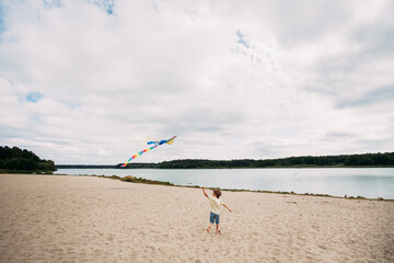 A young cute boy with a kite in his hands runs along the sandy beach on the banks of a picturesque river. The guy is wearing denim shorts and a yellow shirt.