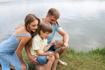 Fototapeta na wymiar A happy family sit on the bank of a picturesque river and play on the tablet together. Mom is wearing a blue dress, dad and son are in denim shorts and t-shirts.
