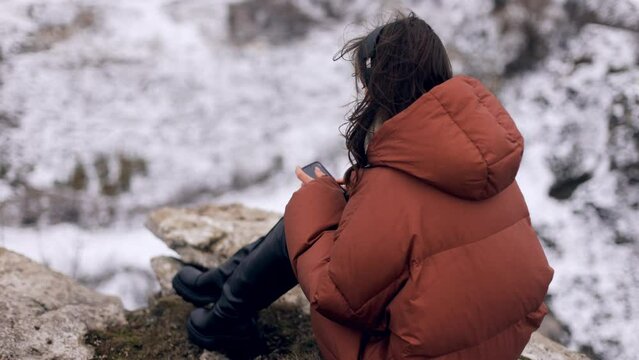 Young attractive beautiful girl woman in winter clothes sitting on the top of a rock with an incredible view and using a smartphone, then looking at the camera and smiling. side view