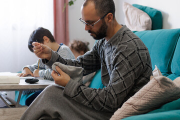 Man sewing a sock in the living room at home while his children do their homework