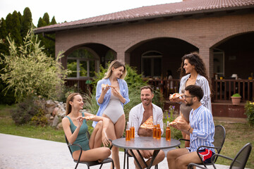 Group of young people cheering with cider and eating pizza by the pool in the garden