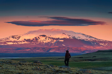 Female adventurer standing on field and volcanic mountain with colorful sky in the dusk among remote wilderness at Highlands of Iceland