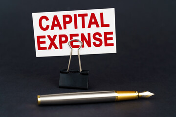 On the black surface is a pen, an office paper clip with a business card on which is written - Capital Expense