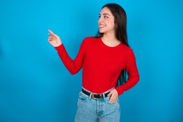 Fototapeta na wymiar Smiling young brunette girl wearing red T-shirt against blue wall indicating finger empty space showing best low prices