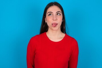 young brunette girl wearing red T-shirt against blue wall showing grimace face crossing eyes and showing tongue. Being funny and crazy