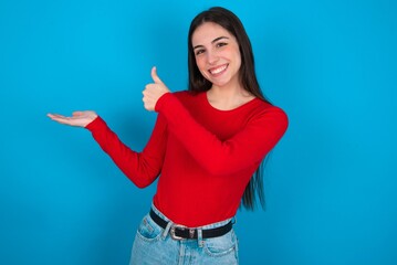 young brunette girl wearing red T-shirt against blue wall Showing palm hand and doing ok gesture with thumbs up, smiling happy and cheerful.