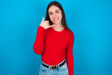 young brunette girl wearing red T-shirt against blue wall smiling doing phone gesture with hand and fingers like talking on the telephone. Communicating concepts.