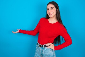 Fototapeta na wymiar young brunette girl wearing red T-shirt against blue wall feeling happy and cheerful, smiling and welcoming you, inviting you in with a friendly gesture