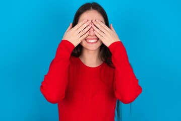 young brunette girl wearing red T-shirt against blue wall covering eyes with hands smiling cheerful and funny. Blind concept.