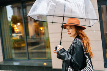 Side view of elegant redhead young woman wearing fashion hat holding transparent umbrella walking...