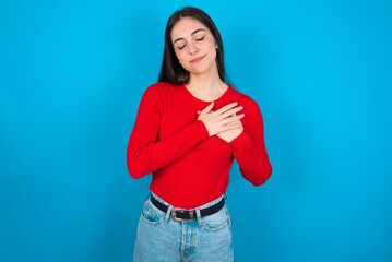 young brunette girl wearing red T-shirt against blue wall smiling with hands on chest with closed...