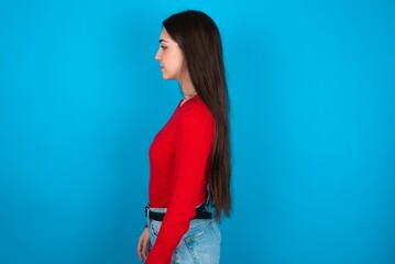 young brunette girl wearing red T-shirt against blue wall looking to side, relax profile pose with natural face with confident smile.