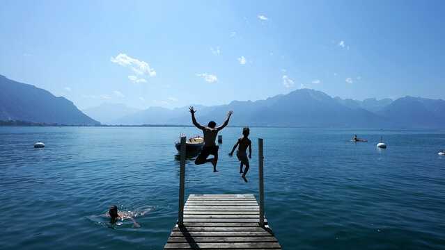 People Jumping Into Lake Water. Father And Son Running In Swiss Pier And Diving Into Geneva Lake Landmark. Parent And Teen Kid Enjoying Holidays