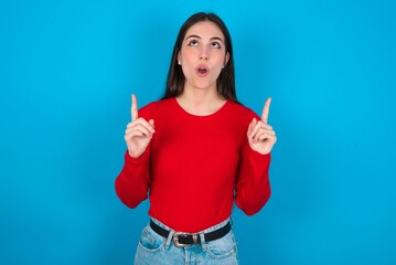 young brunette girl wearing red T-shirt against blue wall amazed and surprised looking up and pointing with fingers and raised arms.