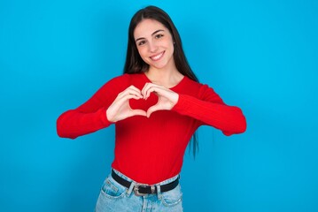young brunette girl wearing red T-shirt against blue wall smiling in love doing heart symbol shape...